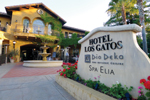 Entrance of Hotel Los Gatos with a stone sign listing Dio Deka restaurant and Spa Elia, surrounded by flowers. The building is yellow with arched windows and a fountain in front.