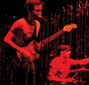 A musician plays an electric guitar and sings into a microphone on stage, while a drummer plays in the background. Red lighting and shiny streamers create a vibrant concert atmosphere.