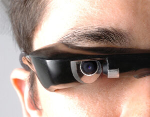 A close-up of a person wearing black smart glasses with a small built-in camera above the right eye, focusing on the eye area and the technology. The background is neutral and out of focus.