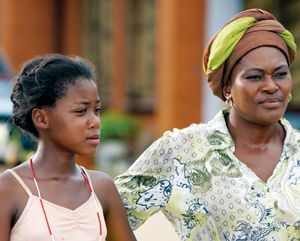 A girl and a woman stand side by side outdoors, both looking intently at something off-camera. The girl wears a light-colored dress, and the woman wears a patterned shirt and a brown and green headwrap.