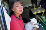 A woman with brown hair in a red shirt sits inside a vehicle, holding a white cat on her lap. A small folding table and various personal items are visible in the background.