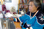 A person wearing a blue San Jose Sharks hockey jersey sits at a desk, typing on a computer keyboard in a cluttered room with bookshelves and a fan in the background.