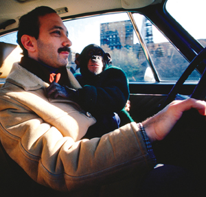 A man with a mustache drives a car while a young chimpanzee in a coat sits beside him, looking out the window. Sunlight comes through the windows, and buildings are visible outside.