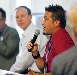 A man in a red shirt and striped tie speaks into a microphone during a panel discussion, while two other men sit beside him listening attentively.