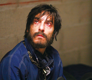 A man with messy dark hair and a beard, wearing a blue shirt and a chain around his neck, sits against a concrete wall, looking up with red, tired eyes and a distressed expression.
