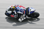 A motorcycle racer in full gear leans sharply into a turn on a racing track, riding a blue and white sports bike at high speed.