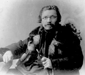 Black and white photo of a man with glasses and a mustache, wearing a fur-trimmed coat and holding a cane, seated and facing the camera with a neutral expression.