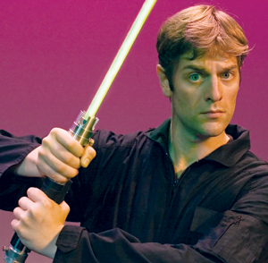 A man in a black outfit holds a glowing green lightsaber with both hands, standing against a pink and purple background, looking intently forward.