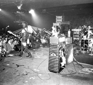 Black and white photo of a rock band performing on stage in front of a large crowd, with various musical equipment and instruments visible. The audience watches closely from in front of the stage.