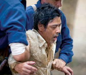 A distressed man with wet hair and a tan shirt is being supported and helped up by two people in blue uniforms. He appears exhausted or in pain.