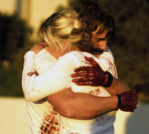 Two people with messy, bloodstained clothes and hands embrace tightly, suggesting strong emotion. Both have light hair, and the background is blurred with trees and a building visible.