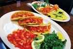 A plate with three hard-shell tacos filled with diced tomatoes and meat, served with sour cream, chopped tomatoes, cilantro, and lemon wedges; another plate with an omelette and vegetables in the background.