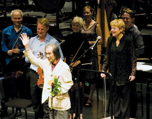 An older man in a light jacket stands on stage holding flowers and waving, with several smiling musicians and a woman in black standing behind him, in an orchestral concert setting.