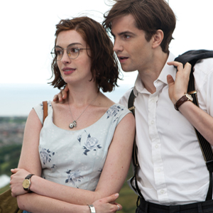A woman with short brown hair and glasses stands next to a man in a white shirt and suspenders, who has his jacket slung over his shoulder. They are outdoors, with a blurred landscape in the background.