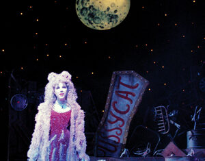 A performer dressed in a sparkly cat costume stands on stage under a large moon backdrop, with “PUSSYCAT” sign and various props resembling junkyard items in the background.