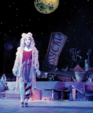 A performer dressed in a sparkly cat costume stands on stage under a large moon backdrop, with “PUSSYCAT” sign and various props resembling junkyard items in the background.