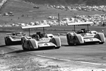 Three race cars with large rear wings speed around a bend on a race track, with a grassy field and parked cars in the background. The image is in black and white, capturing a vintage motorsport scene.