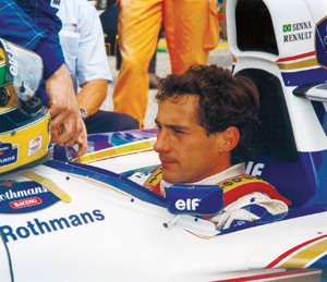 A race car driver sits in a Formula 1 car with “Rothmans” and “elf” logos, while a person hands him a helmet. The driver looks focused, wearing a racing suit. The scene is at a racetrack.