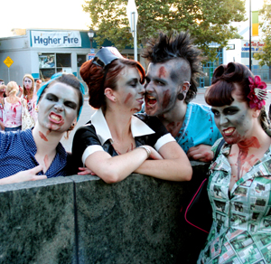 Four people in zombie makeup and vintage-style clothing pose together outdoors, snarling and baring their teeth. A crowd of similarly dressed people is visible in the background, suggesting a zombie-themed event or parade.