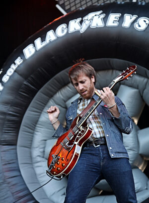 A musician in denim clothing passionately plays an electric guitar on stage in front of a large inflatable tire with “Black Keys” written on it.