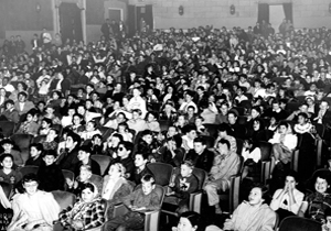 Black-and-white photo of a large audience seated in a theater, with people of various ages facing forward, filling nearly every seat and appearing attentive or engaged.