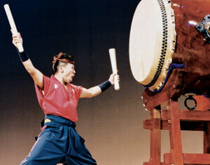 A person in traditional attire energetically plays a large Japanese taiko drum with wooden sticks, captured mid-performance on stage.