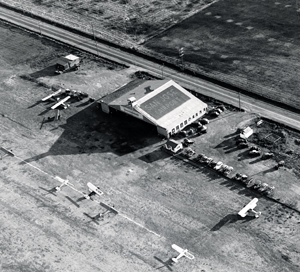 Aerial view of a small airfield with several vintage airplanes parked near a hangar. Cars are lined up in front of the building, and a road runs parallel to the airfield in the background.