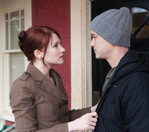 A woman with red hair and a brown coat holds onto the front of a mans black jacket as they stand face to face outside a building; the man wears a gray knit beanie and looks serious.