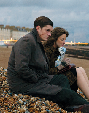 A man and a woman sit on a rocky beach at dusk, both wearing coats. The man looks ahead with a serious expression, while the woman, holding a lace handkerchief, gazes downward. Buildings and lights are visible in the background.