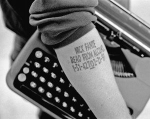 A person’s forearm with the text “Mick Fante Dead From Alcohol 1-31-42 (72-21-91)” tattooed on it, resting on a typewriter.