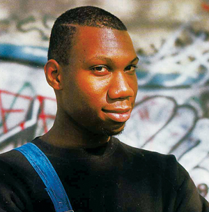 A man with closely cropped hair and a trimmed beard wears a black shirt and blue overalls, standing in front of a graffiti-covered wall, looking confidently at the camera.