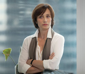 A woman with short brown hair, wearing a white blouse and gray scarf, stands indoors with her arms crossed, looking to the side with a serious expression. A blurred plant and cityscape are in the background.