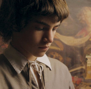 A young person with short brown hair and a white-collared shirt looks down thoughtfully, standing in front of a blurred, classical painting background.
