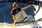 A skateboarder wearing a helmet and casual clothes is performing a trick on the curved edge of a skatepark bowl, with an audience watching in the background.