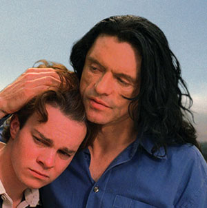 Two men stand close together outdoors. The man on the right with long black hair is gently holding and comforting the younger man on the left, who has short brown hair and looks sad.