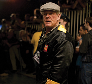 A man wearing a flat cap and a black satin jacket with yellow trim stands in a dimly lit room, looking over his shoulder. There are blurred people and metal bars in the background.