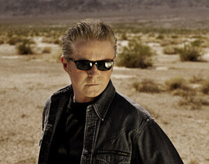 A man wearing sunglasses, a dark shirt, and a denim jacket stands in a dry, desert landscape with sparse bushes and distant mountains under a cloudy sky.