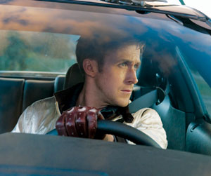 A man wearing a white jacket and brown driving gloves sits behind the wheel of a car, looking intently out the side window with both hands on the steering wheel.
