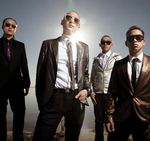 Four men in suits and sunglasses stand outdoors in sunlight, with water and sky in the background. They pose confidently, with the sun shining behind them, casting a dramatic, stylish effect.