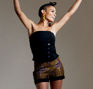 A smiling woman with braided hair poses with arms raised, wearing a black strapless top and patterned shorts with yellow and brown designs, along with yellow earrings.