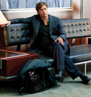 A man with light brown hair sits on a black leather couch, dressed in a dark blazer, green shirt, and dark pants. He has a serious expression, with one arm resting on the back of the couch. A bag and coat sit in front of him.