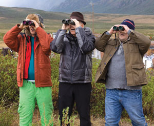Three people in winter clothing stand outdoors in a grassy area, each looking through binoculars toward the distance. Mountains and cloudy skies are visible in the background.