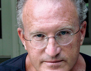 A middle-aged man with short gray hair, wearing round glasses and a black shirt, looks directly at the camera with a serious expression.