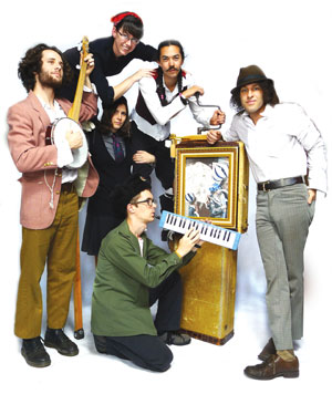 Six people pose together with vintage musical instruments, including a banjo and a small upright piano. They wear eclectic, retro-inspired clothing and stand against a plain white background.
