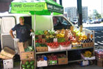 A person stands beside a NYC Green Cart filled with fresh fruits and vegetables, parked on a city street with a white van in the background. Various produce items are displayed in crates and boxes.