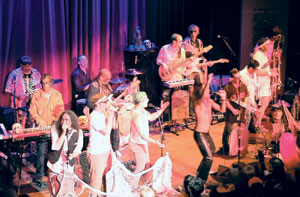 A lively band performs on stage with multiple musicians playing instruments, including guitars, keyboard, and brass, under colorful stage lights. The audience watches as one performer dances energetically in the center.