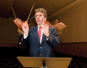 A conductor in a suit stands at a music stand, moving his baton with blurred motion, indicating dynamic arm movement. The background shows empty seats in an auditorium.