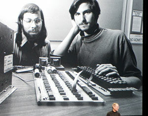 A man stands on stage in front of a large projected black-and-white photo of two young men with early computer hardware.