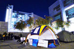 A small tent with signs attached is set up on a city sidewalk at night, surrounded by a few trees and nearby buildings illuminated by lights in the background.