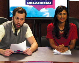 Two news anchors sit at a desk with papers, presenting on a show called Good News Oklahoma! A man in a gray vest and a woman in a red top smile in front of a TV screen with the show’s name.